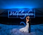 bride and groom stand in front of blue neon sign that reads "We belong here"